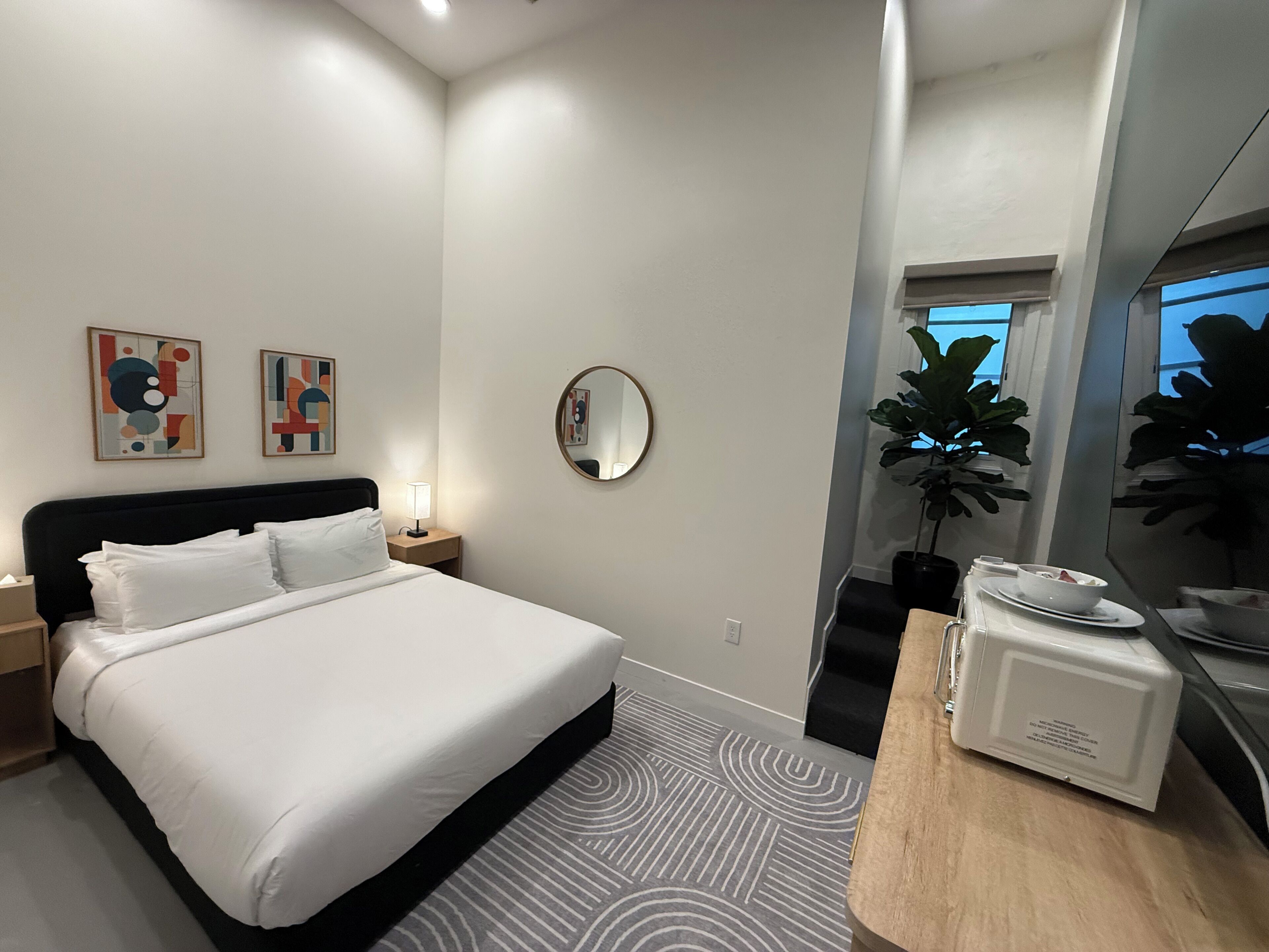 A modern hotel room with a bed, bedside table, lamp, artwork, mirror, potted plant, and a microwave.