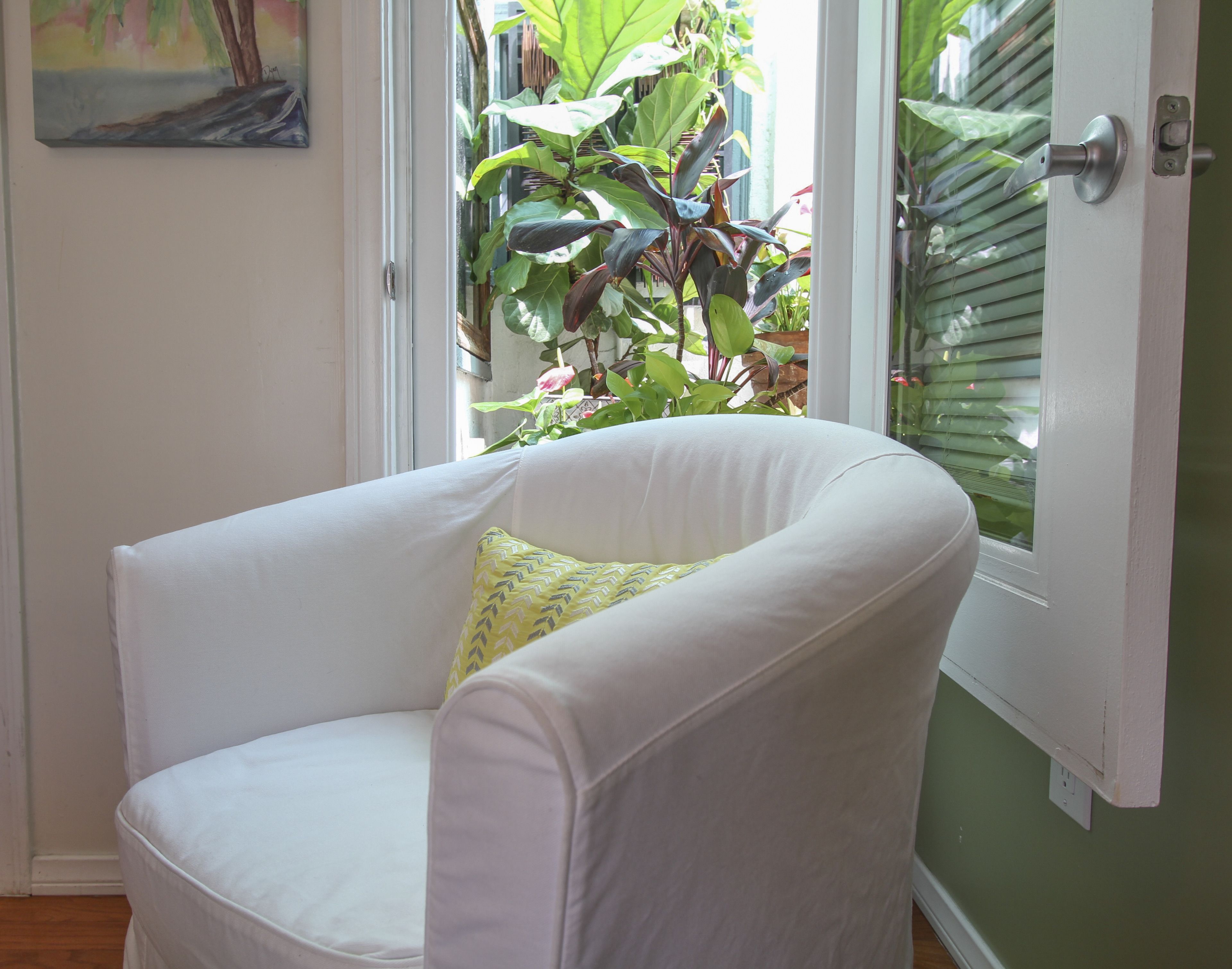 A white armchair with a yellow pillow in a room with a green wall and a window with blinds.