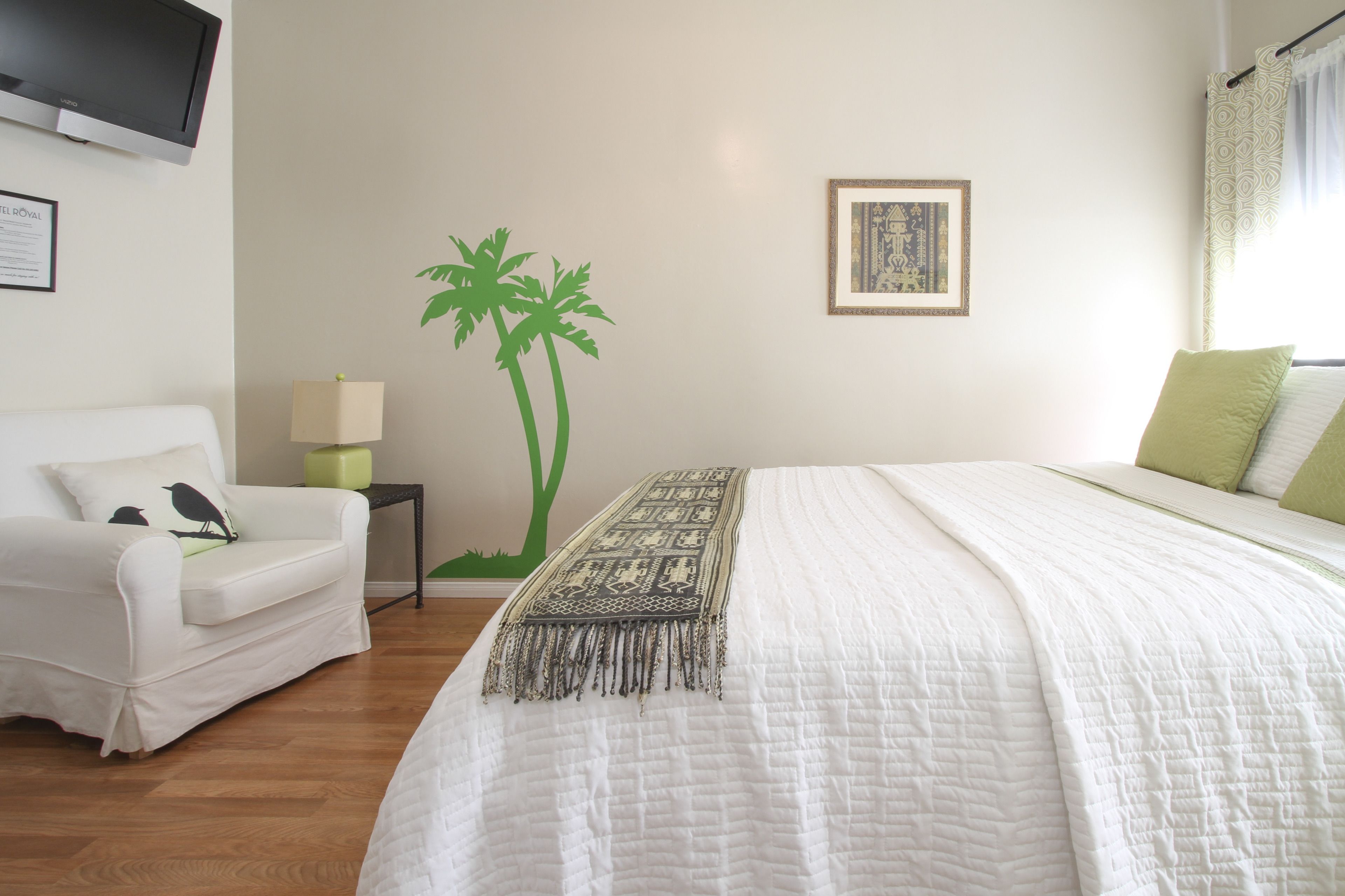 A hotel room with a bed, a white armchair, a small table with a lamp, a wall-mounted TV, and a framed picture.