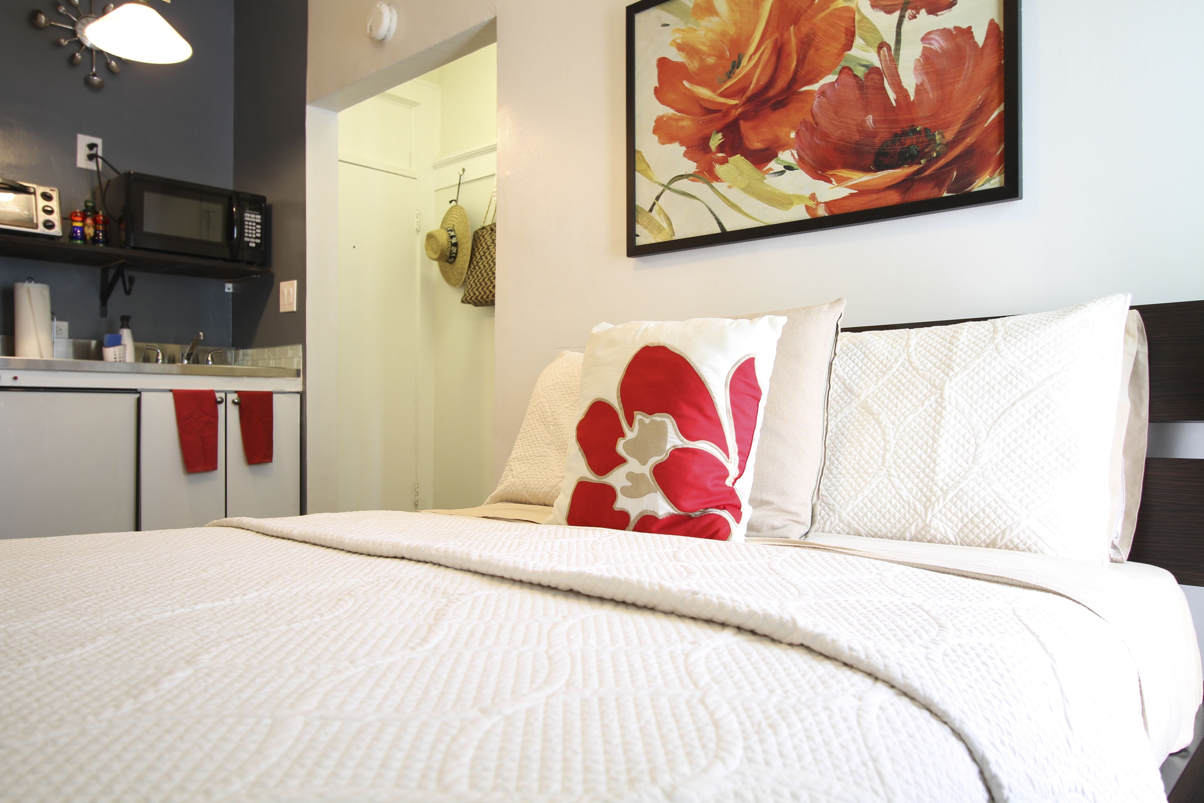A neatly made bed with a white and red patterned blanket, a microwave above the counter, and a floral painting on the wall.