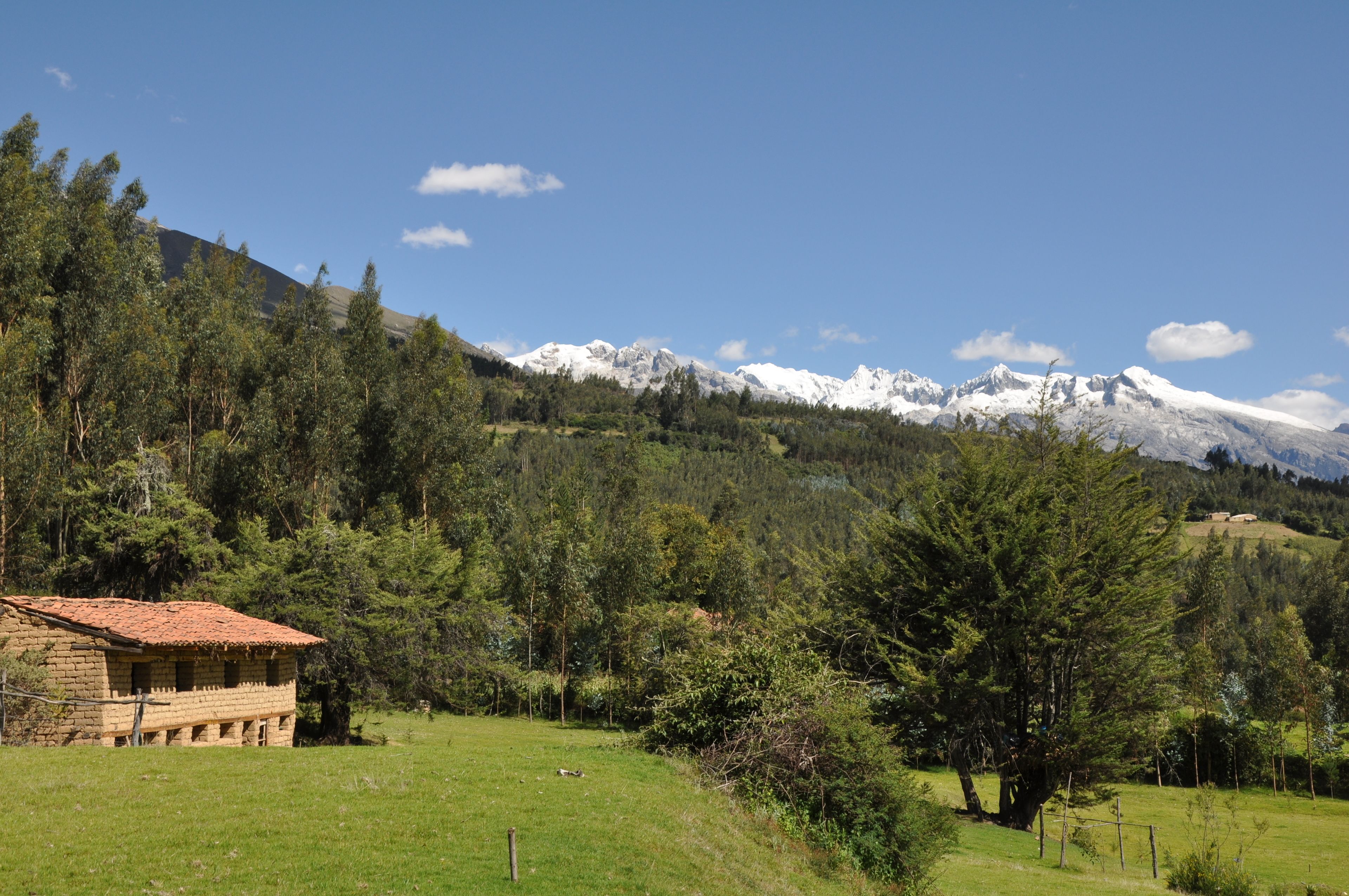 Um edifício rústico de pedra com telhado de telhas vermelhas, rodeado por uma paisagem verde exuberante e ao fundo montanhas nevadas sob um céu azul límpido.