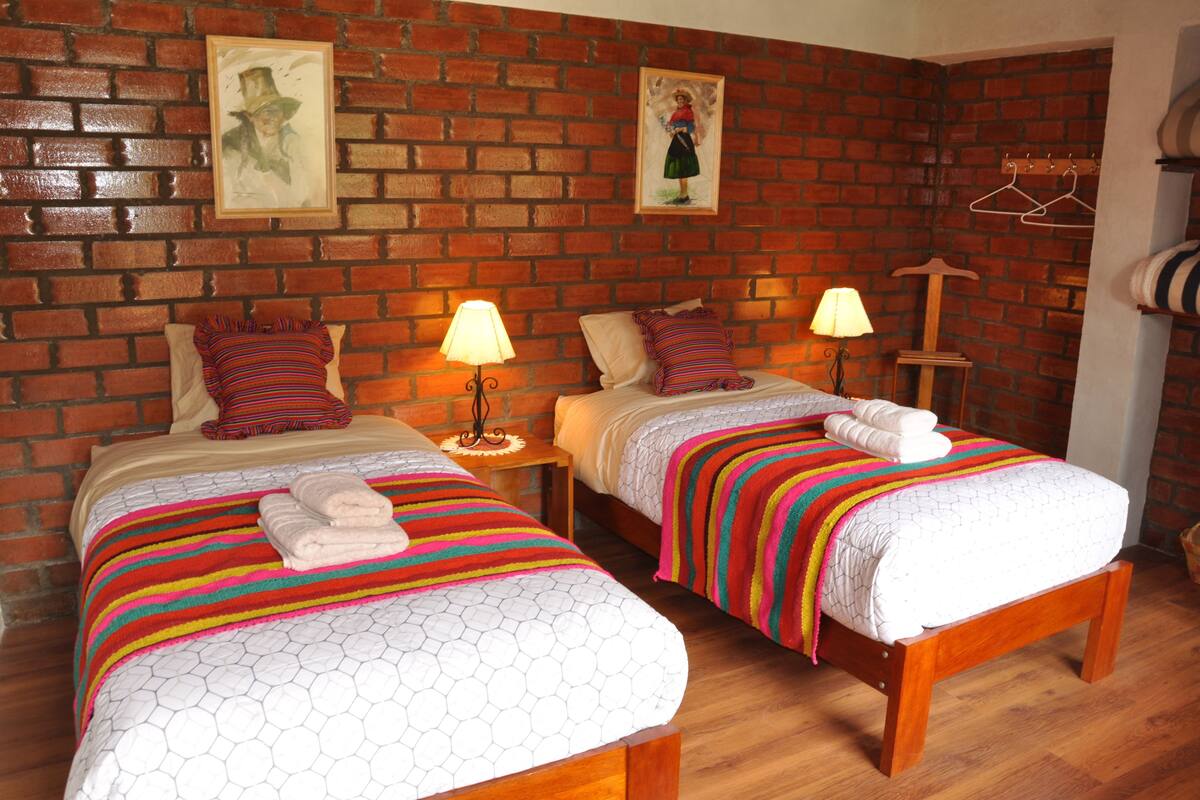 Quarto com duas camas, cada uma com um cobertor e almofadas listradas e coloridas, uma parede de tijolos e piso de madeira.