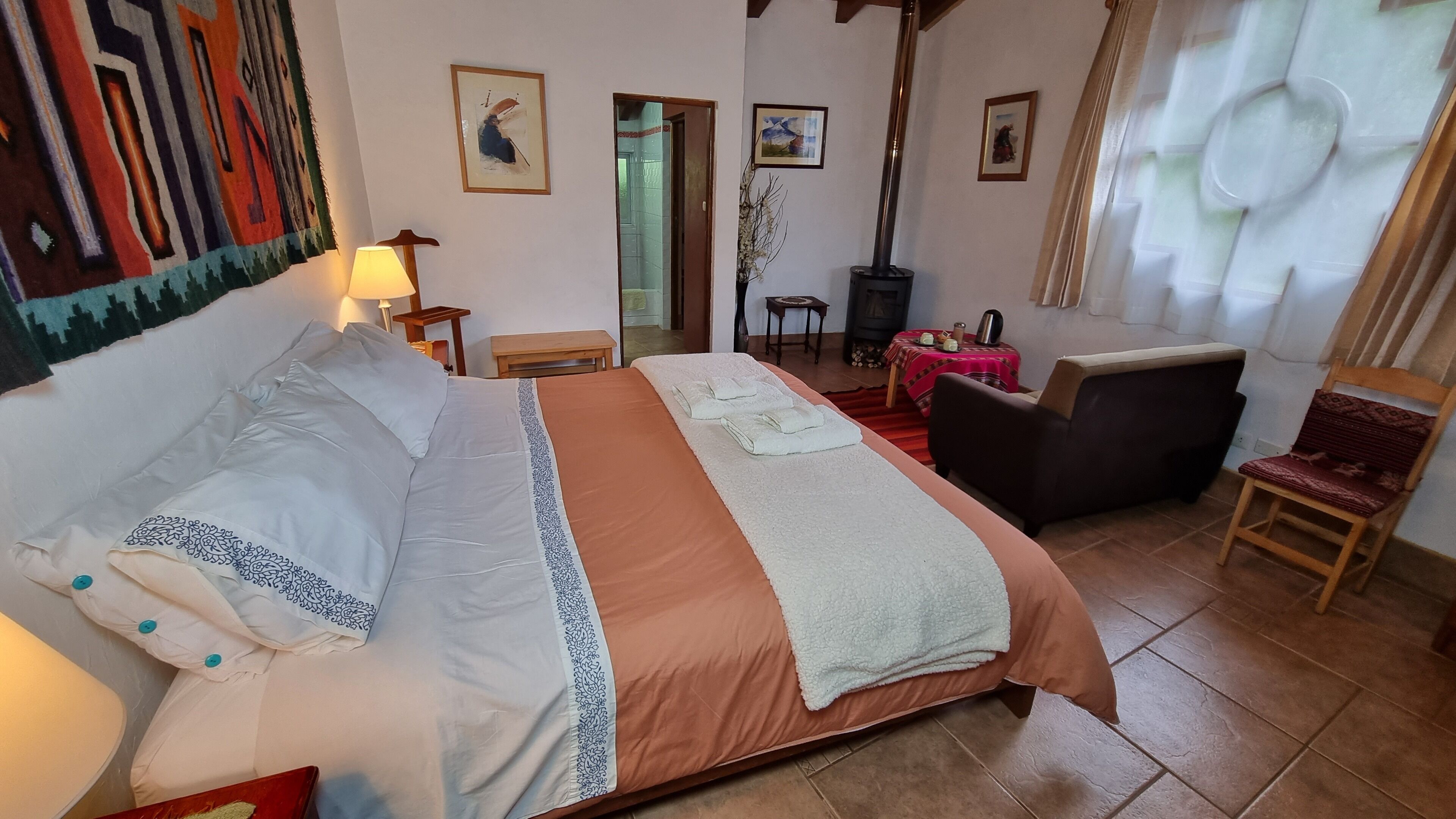 Habitación de hotel con una cama grande, un cabecero de madera, una mesita de noche con una lámpara, una silla, una chimenea y vistas al pasillo.