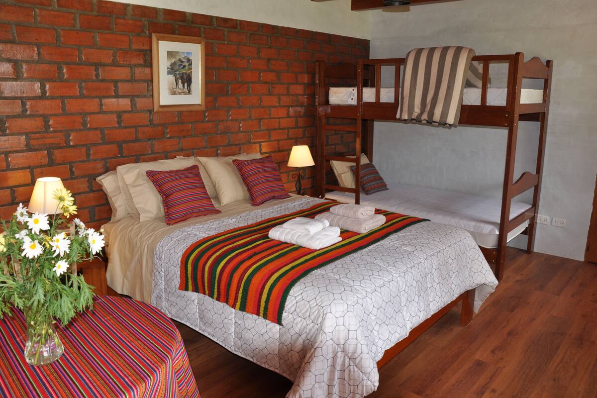 Quarto com parede de tijolos, uma cama de solteiro, cama com roupa de cama listrada, mesa de cabeceira com abajur e um vaso com flores.