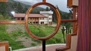 A view through a window with a wooden frame and a circular mirror, overlooking a building with a red roof and a courtyard with cacti and a white car. - Copacabaña Lodge (Carhuaz)
