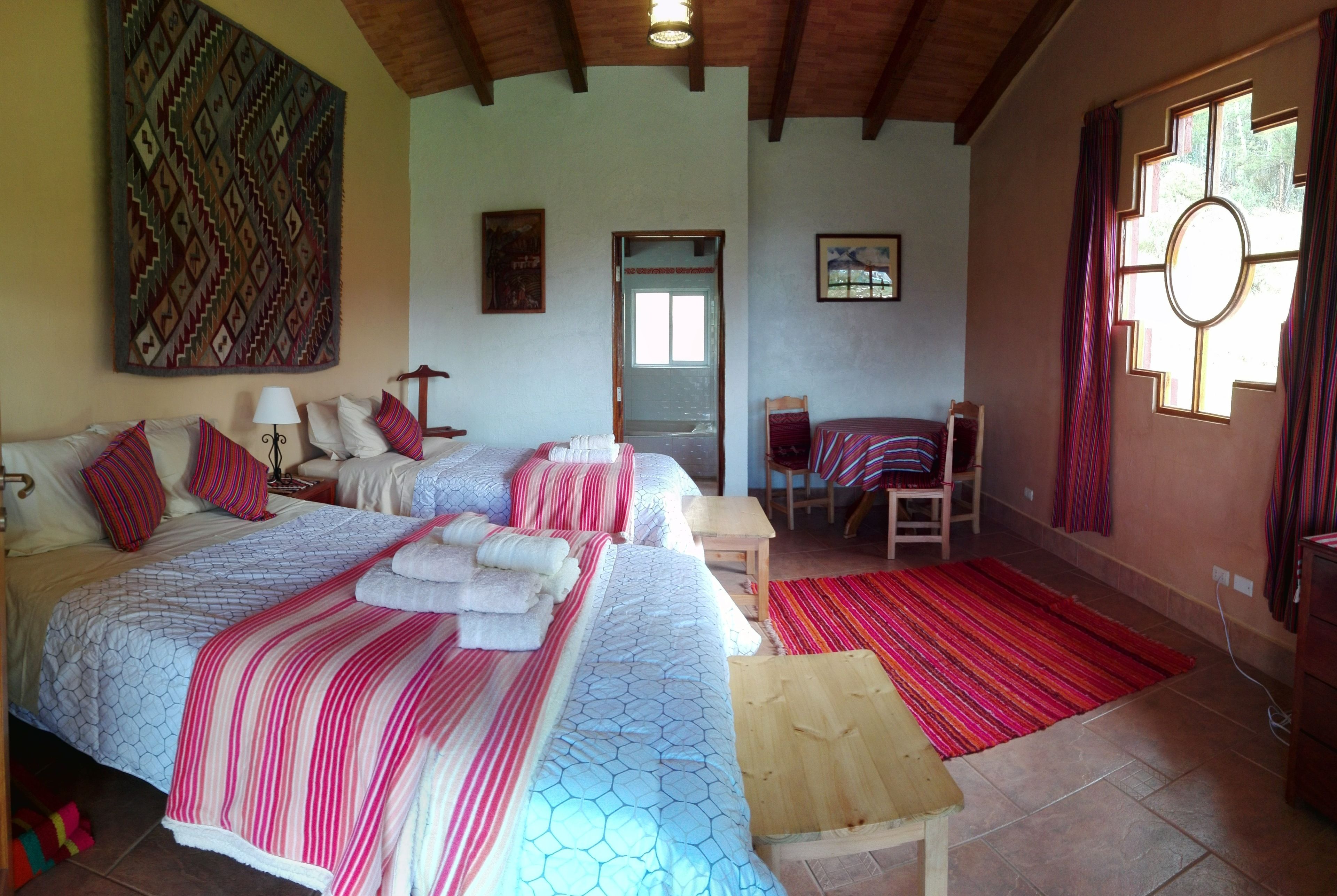 A bedroom with two beds, a wooden bench, a chair, a table, and a large window with curtains.