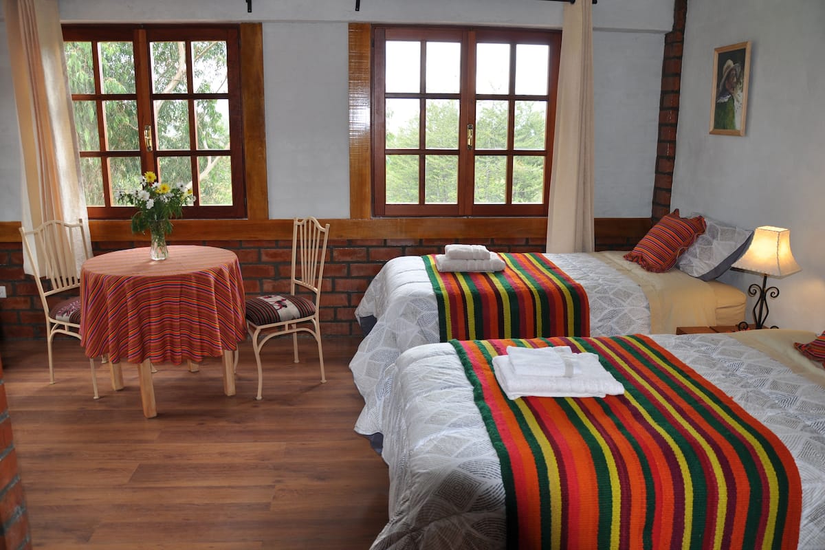 Quarto com duas camas, uma mesa com cadeira e janela com vista para árvores.
