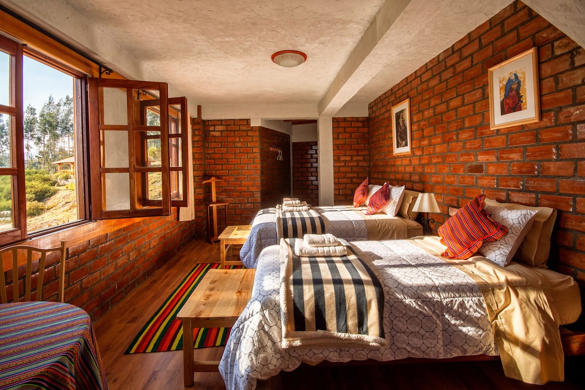 Quarto com duas camas, um banco de madeira, um tapete listrado e colorido, e uma parede de tijolos com quadros emoldurados.