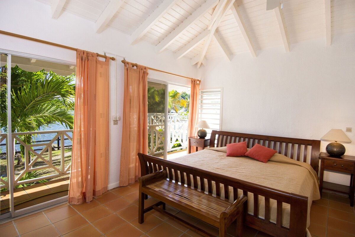 A bedroom with a bed, a bench, a nightstand, and a view of a balcony with plants.