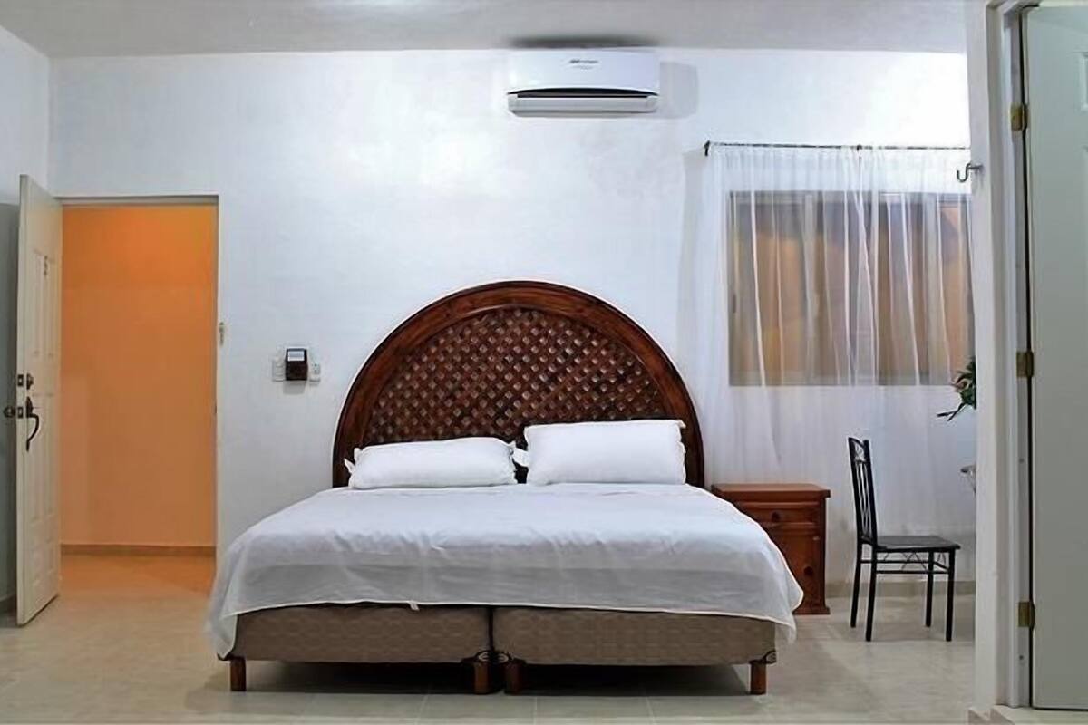 Een slaapkamer met een houten hoofdbord, een bed met wit beddengoed, een nachtkastje, een stoel en een airconditioning.