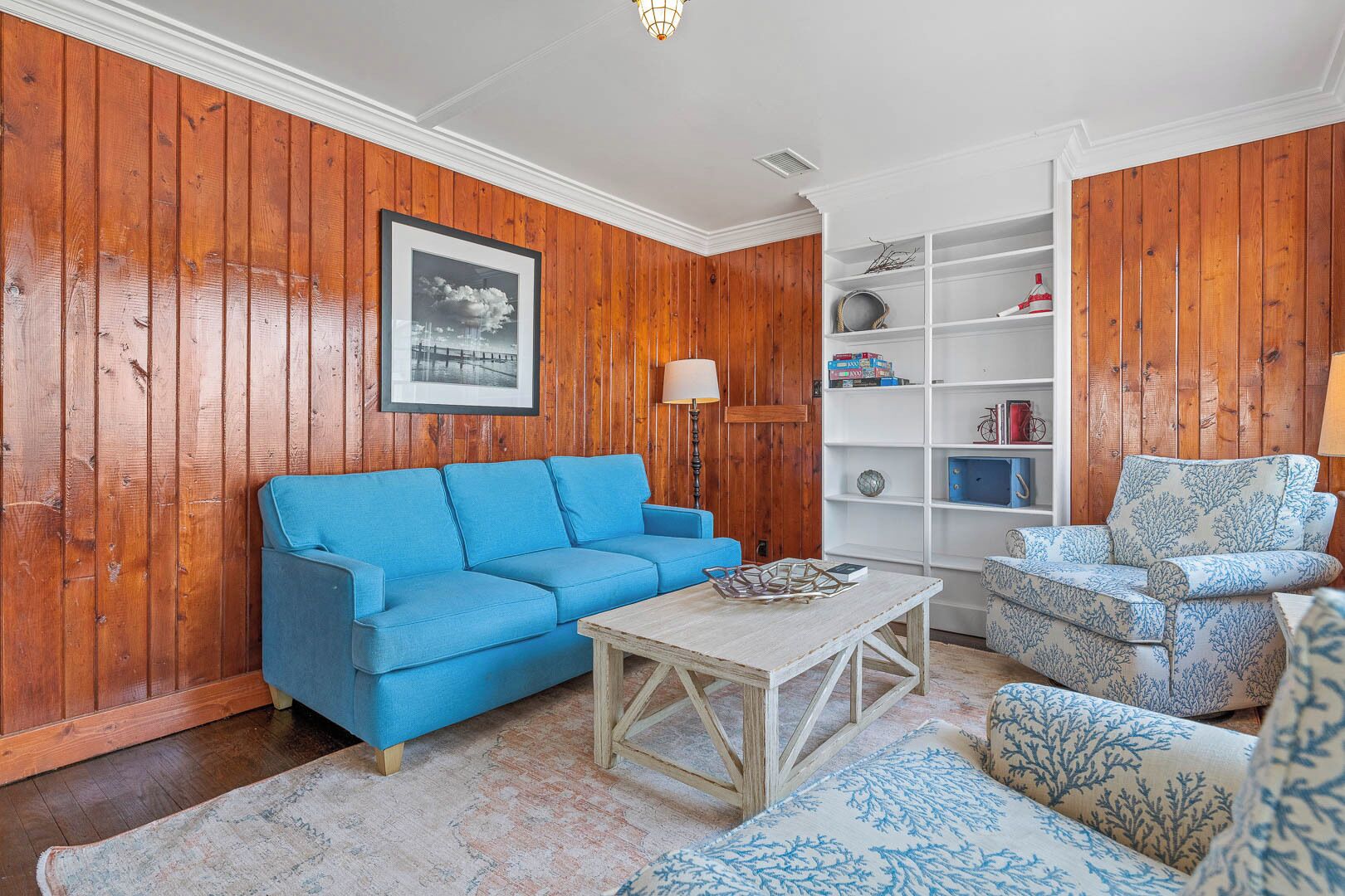 A living room with wooden paneling, a blue sofa, a patterned armchair, a white coffee table, and a bookshelf.