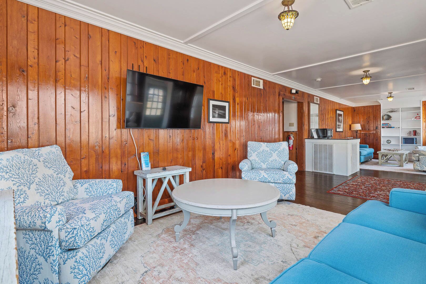 Una sala de estar con paneles de madera, un televisor de pantalla plana, una mesa redonda y sofás azules con estampado.