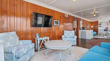 Una sala de estar con paneles de madera, un televisor de pantalla plana, una mesa redonda y sofás azules con estampado.