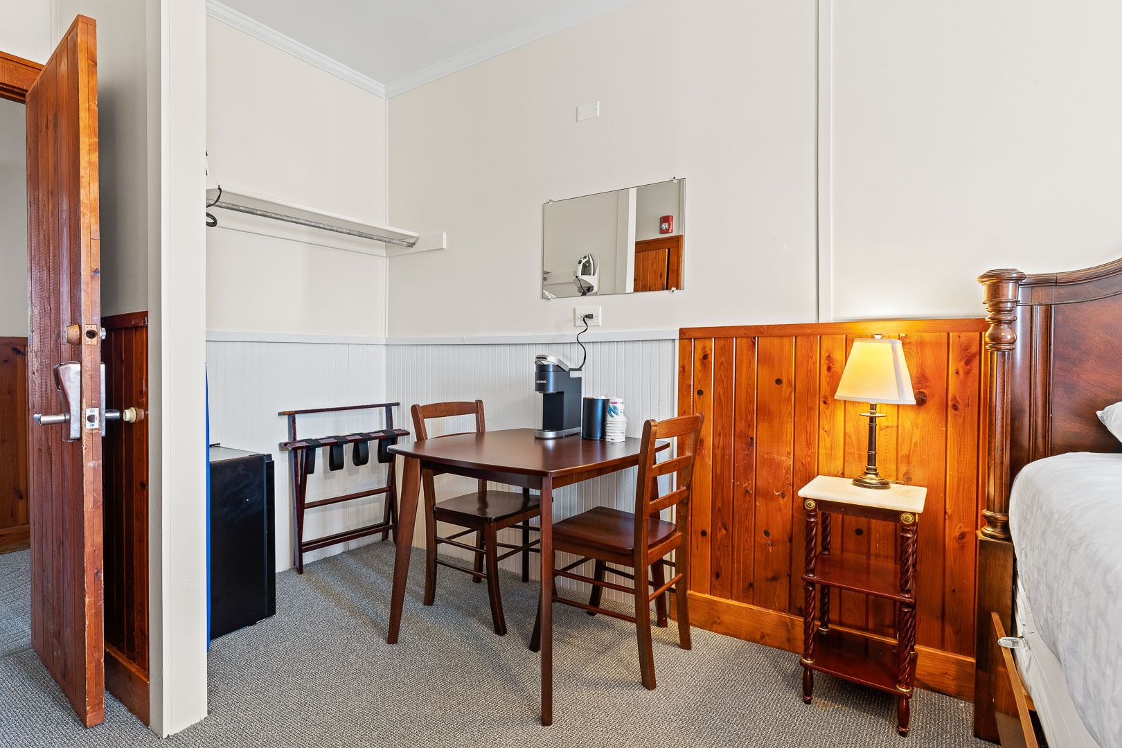 A compact hotel room with a wooden bed, a small desk with a chair, a bedside table with a lamp, a mirror, and a coffee machine.