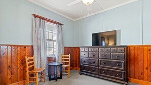 A room with a wooden paneled wall, a dark wooden dresser with multiple drawers, a round table with two chairs, and a window with light blue curtains.