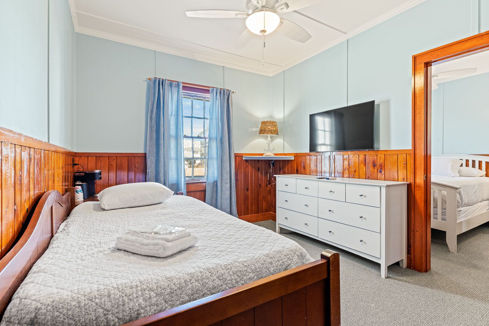 A bedroom with a wooden bed, a white dresser, a flat-screen TV, and a window with blue curtains.