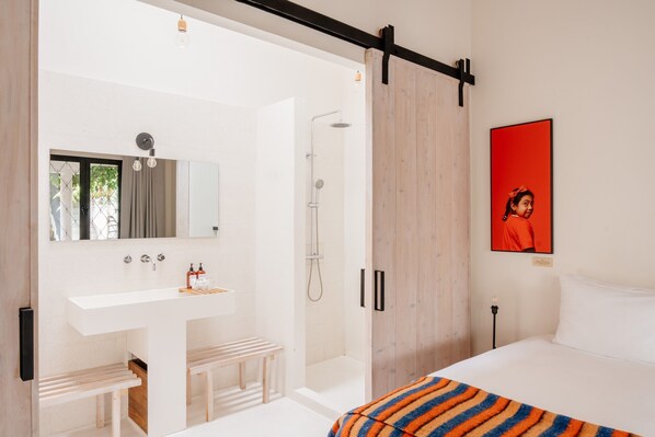 Un baño moderno con un lavamanos blanco, espejo y ducha. Hay un banco debajo del lavamanos y una cama con una manta a rayas de colores.