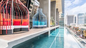 Un hotel moderno con zona de piscina, mobiliario exterior con cojines rojos y azules, y vistas al horizonte de la ciudad.