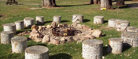 A campfire surrounded by birch logs, with trees and cabins in the background.