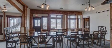 A spacious dining area with multiple tables and chairs, wooden beams on the ceiling, and large windows providing natural light.