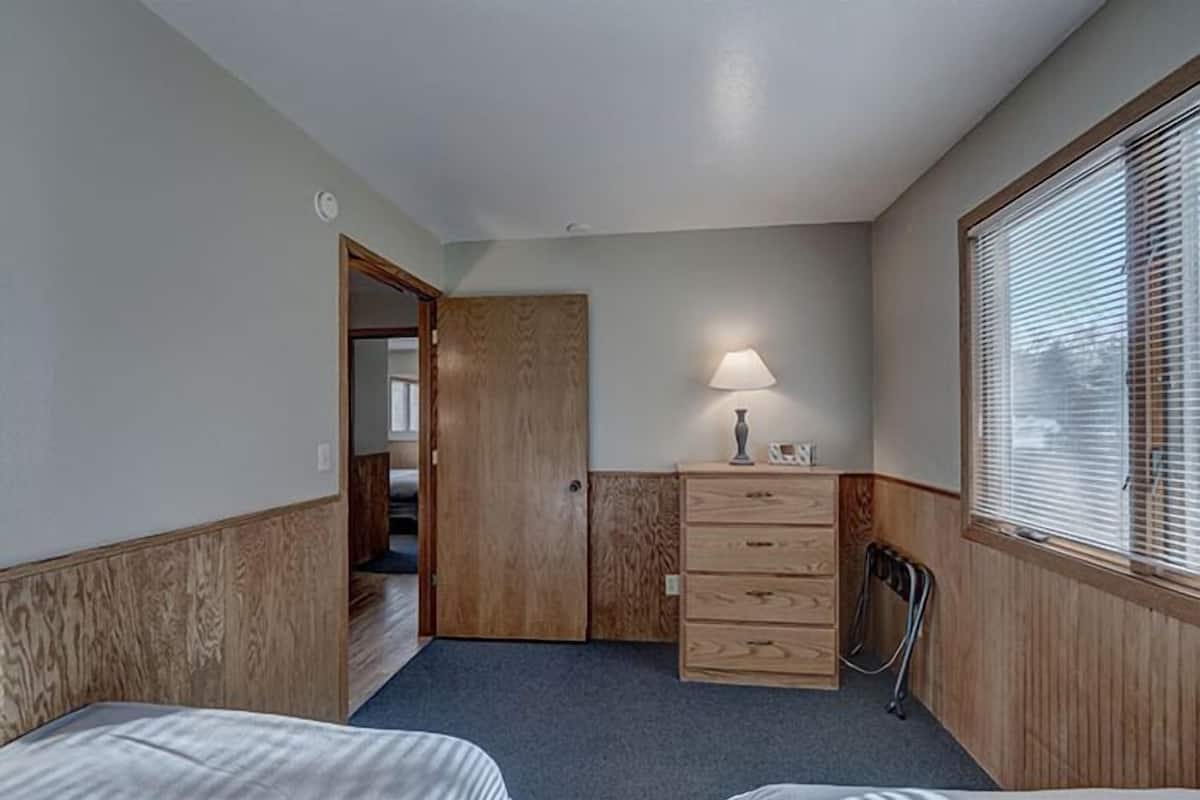 Une chambre de taille modeste, équipée de deux lits, d’une commode en bois, d’une lampe et d’une fenêtre avec des stores.