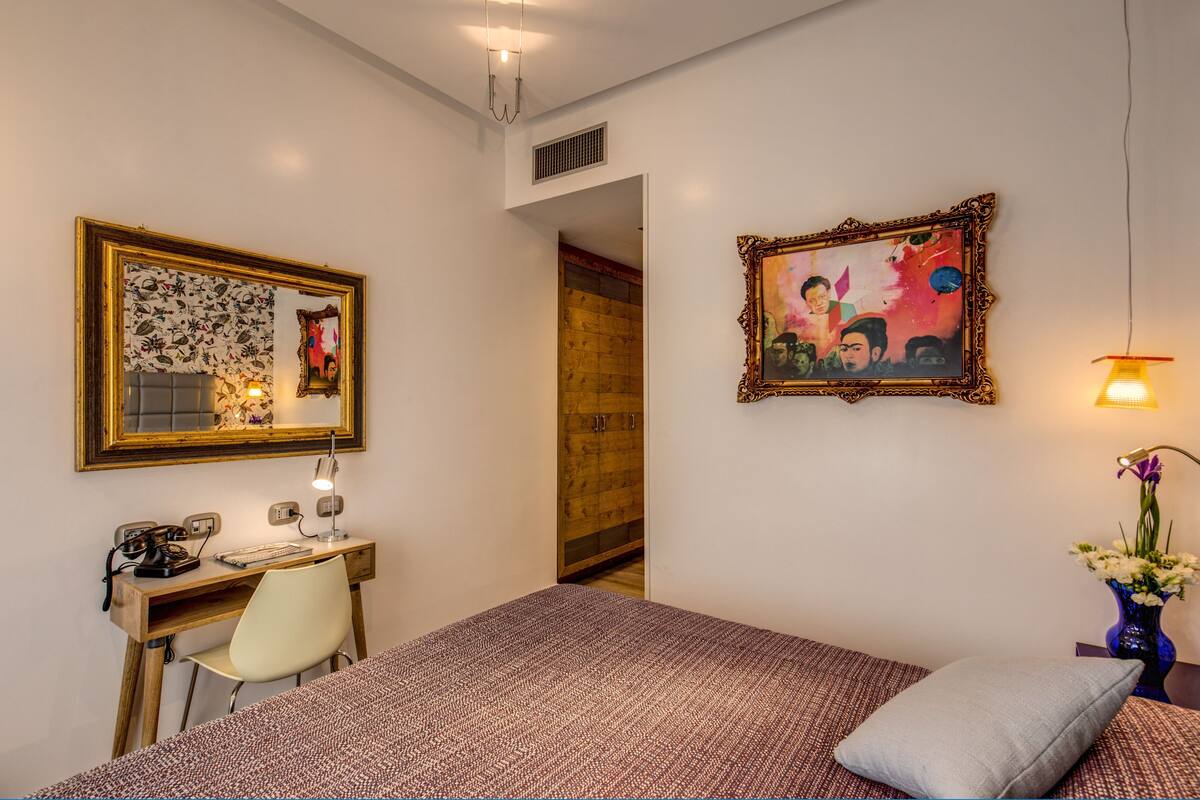A bedroom with a bed, a desk with a chair, a mirror, and a painting on the wall.