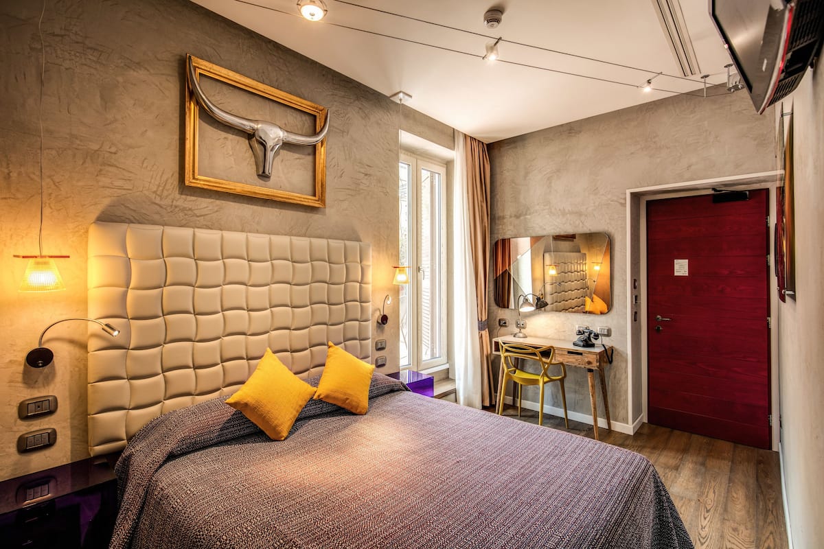 A modern hotel room with a tufted headboard, a desk with a chair, a red door, and a large mirror.