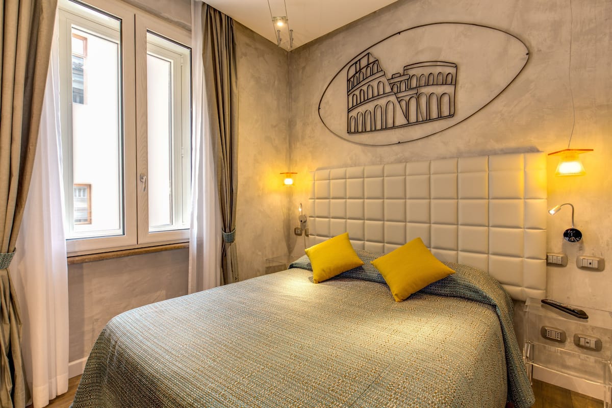 A hotel room with a bed, curtains, a wall-mounted light, and a wall decoration of the Colosseum.