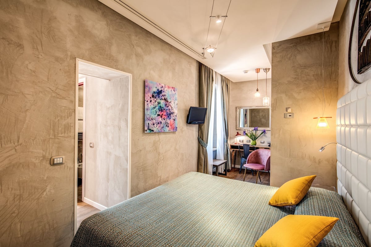 A hotel room with a bed, two pillows, a desk with a chair, a TV, and a bathroom visible through an open door.