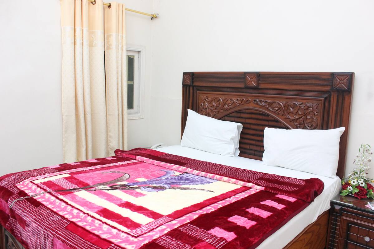 Una cama bien hecha con un edredón rojo y blanco, un cabecero de madera y una mesita de noche con un florero.