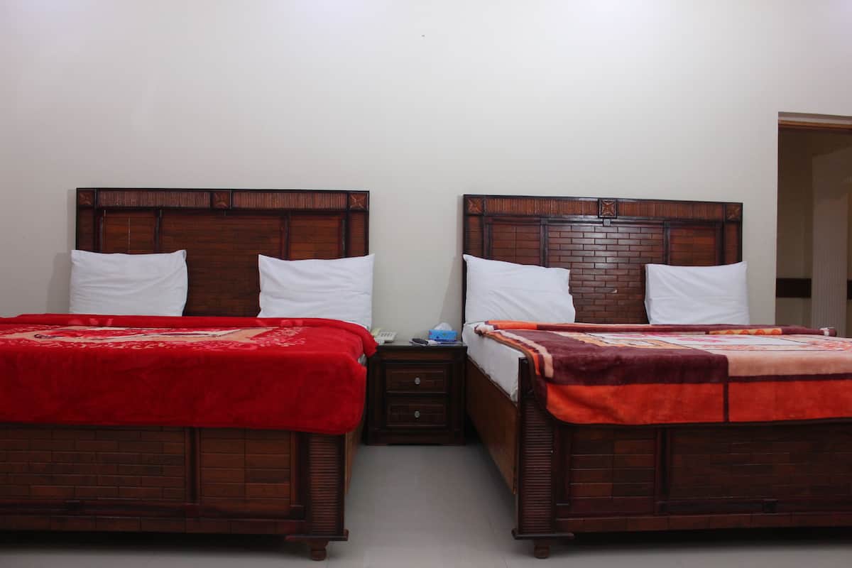 Dos camas de madera con ropa de cama roja y naranja, una mesita de noche de madera con un teléfono y pañuelos, y una pared blanca lisa.