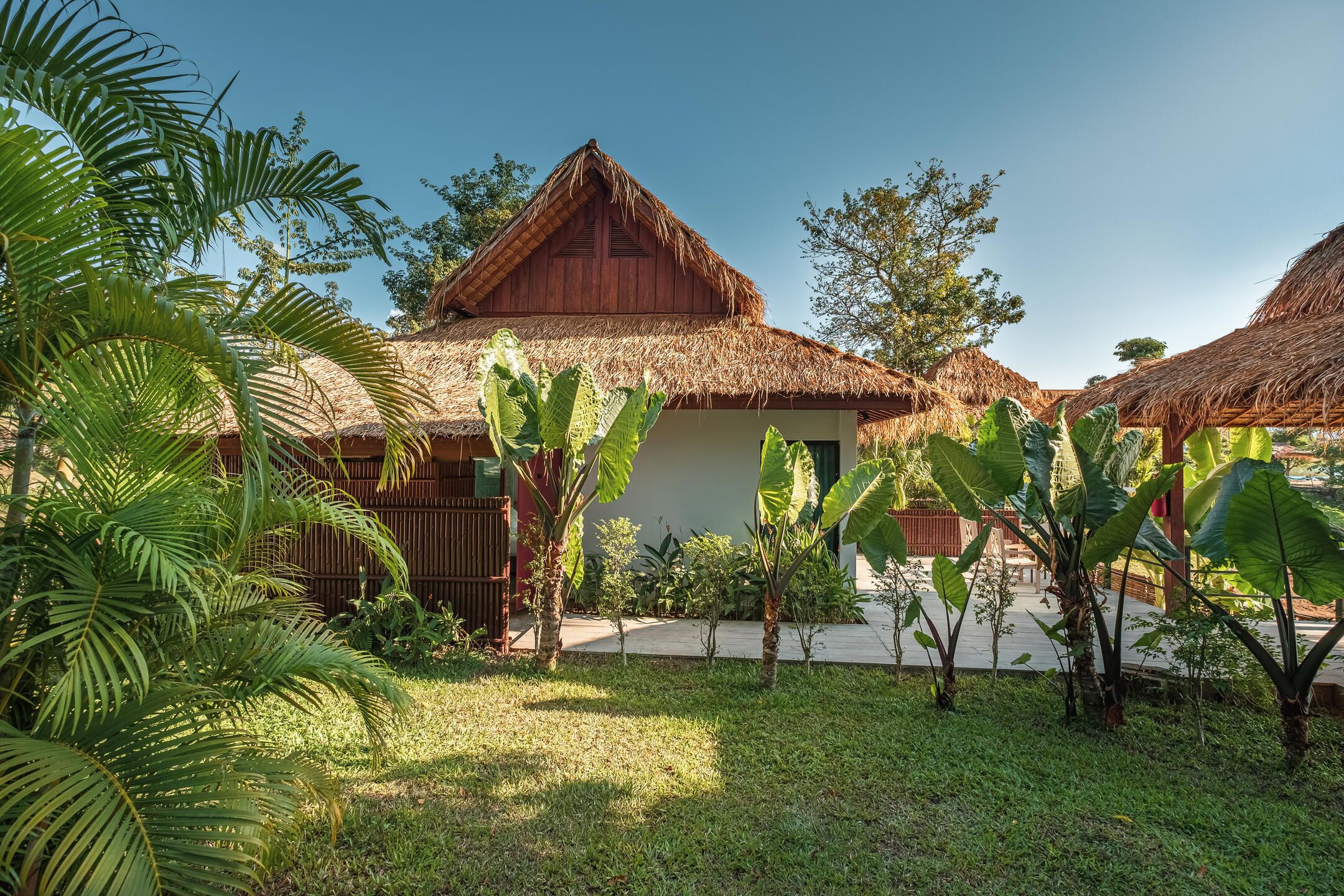 A bungalow with a thatched roof, surrounded by lush greenery and a wooden fence.