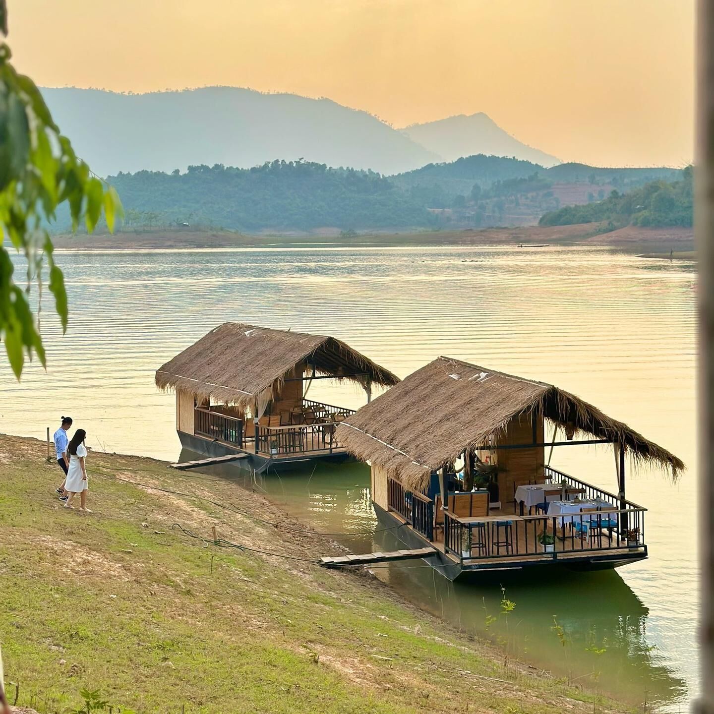 A riverside bungalow with a thatched roof, a wooden deck, and a view of the river and distant hills.