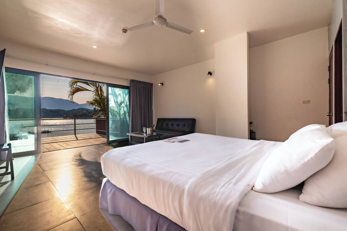 Una habitación de hotel moderna con una cama grande, ventilador de techo, balcón con vistas a las montañas y zona de comedor con mesa y sillas.