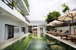 A modern indoor pool area with lounge chairs and umbrellas, surrounded by greenery and a white building facade. - Sam So Boutique Villa (Siem Reap)
