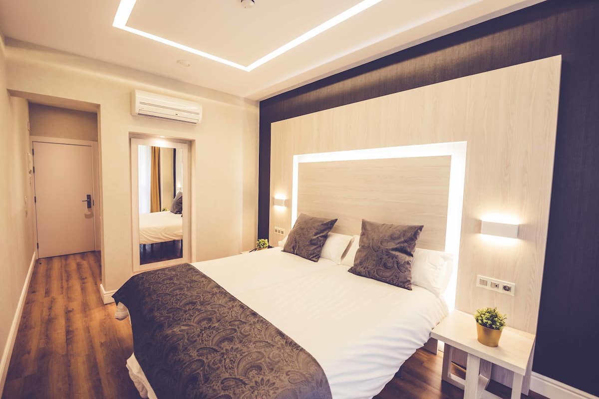 A modern hotel room with a large bed, bedside tables, an air conditioning unit, and a small potted plant.