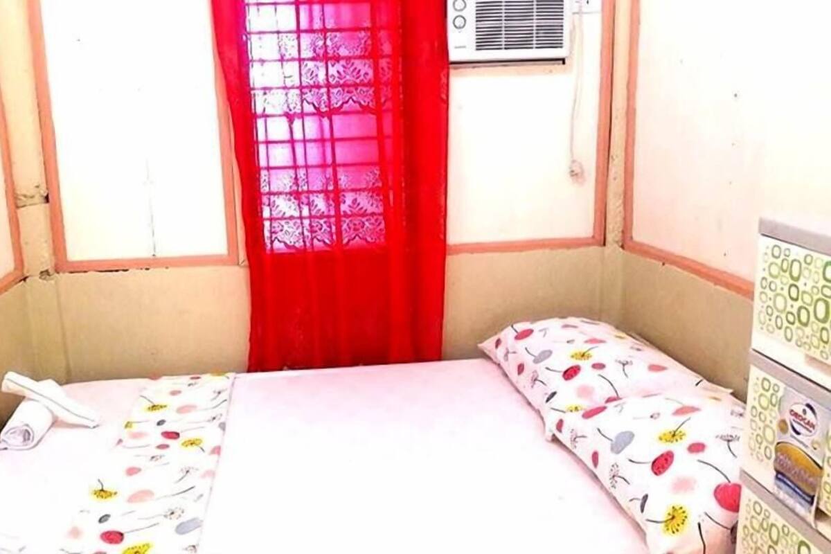 A small, single-bed room with a window, a heater, and a bedside table with a lamp.