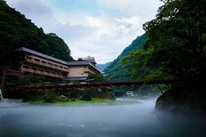 A traditional building with a flat roof and multiple stories, surrounded by lush greenery and a misty river.