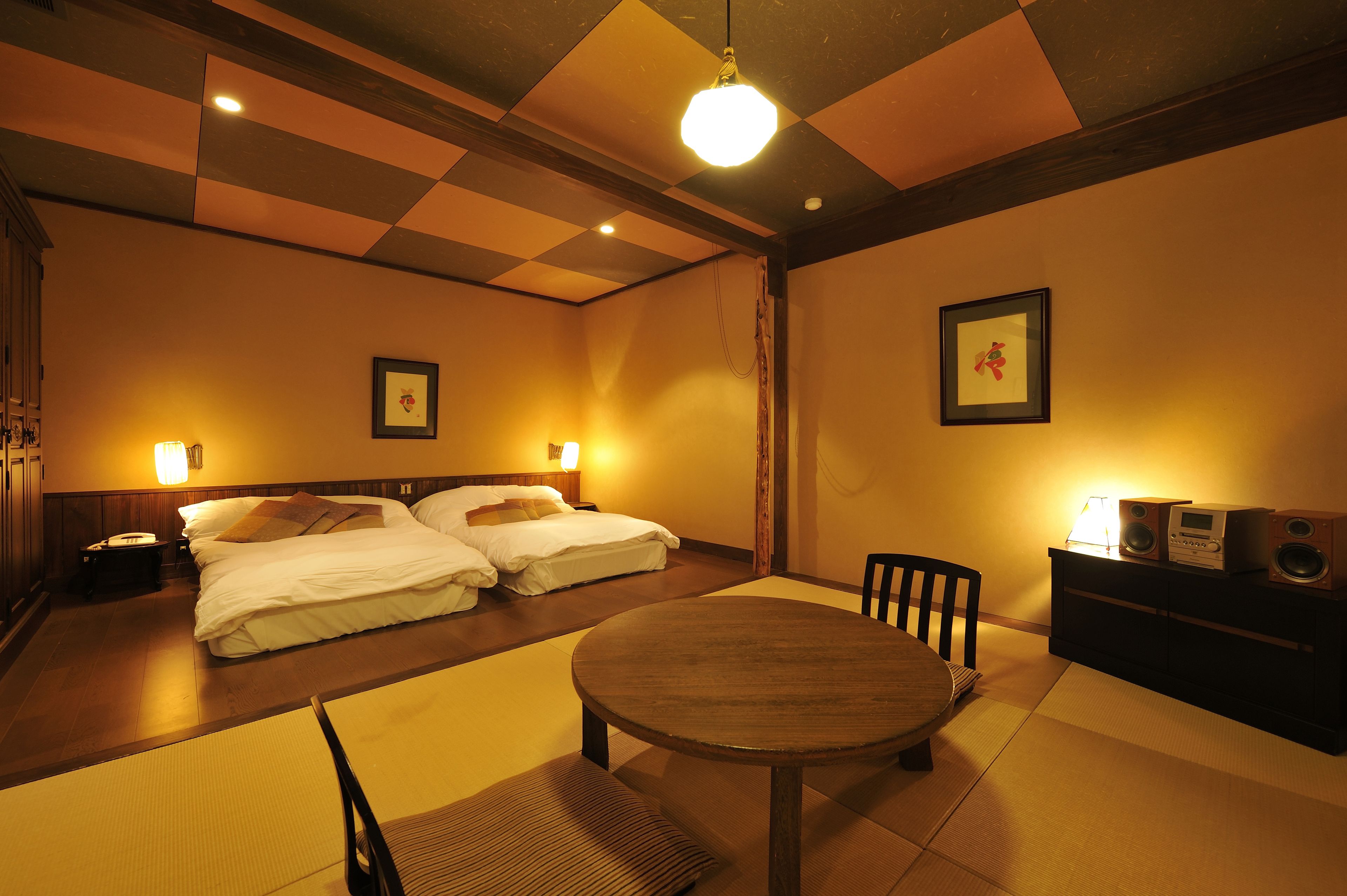 A hotel room with a checkered ceiling, a round wooden table, two beds, a bedside table with a lamp, a chair, and a cabinet with speakers.