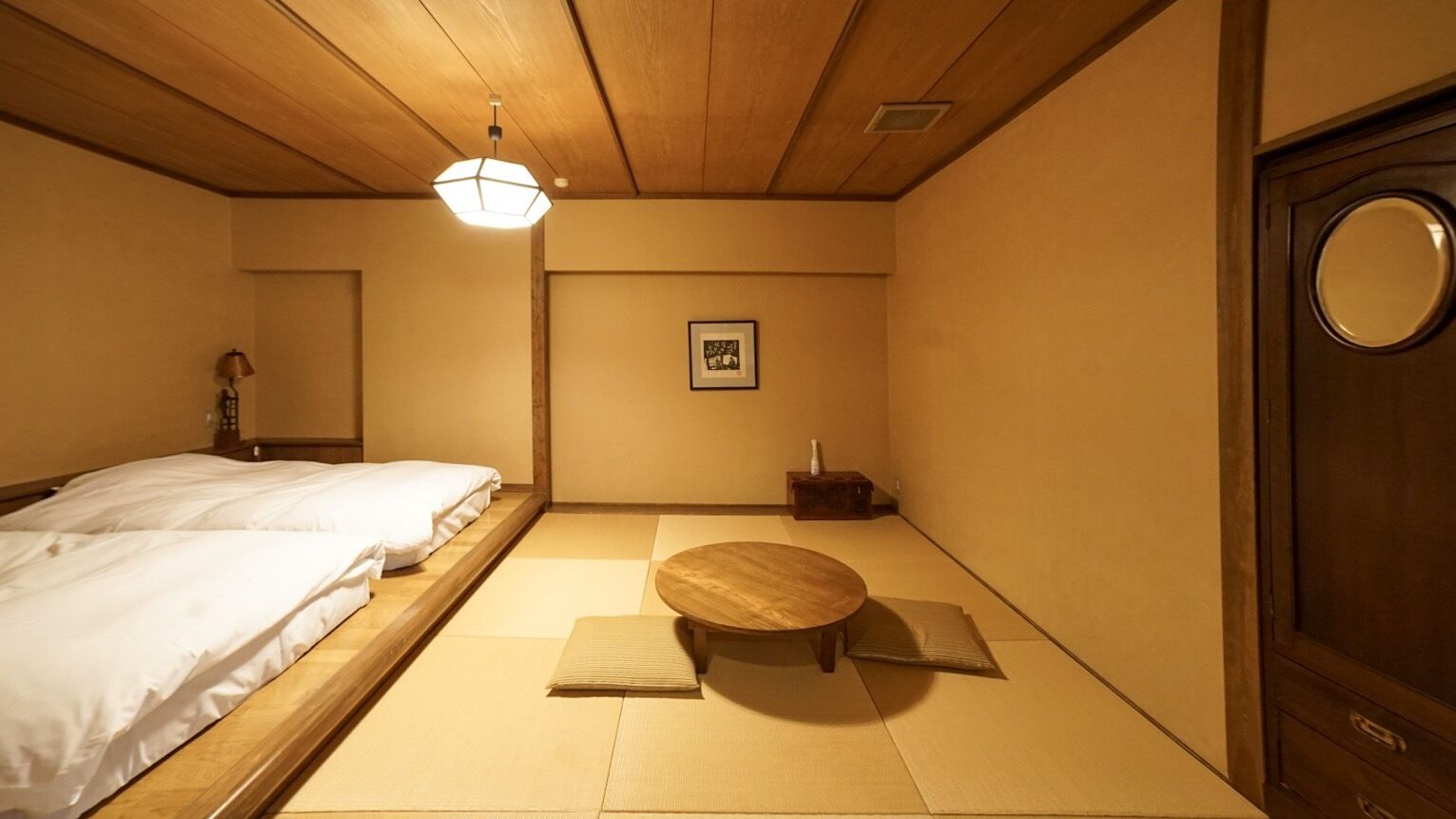 Una habitación japonesa tradicional con suelo de tatami, una mesa de madera baja, una cama con ropa blanca y una puerta de madera con ventanal circular.