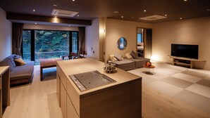 Uma sala de estar moderna com ilha de cozinha, sofá, televisão e uma grande janela com vista para árvores.