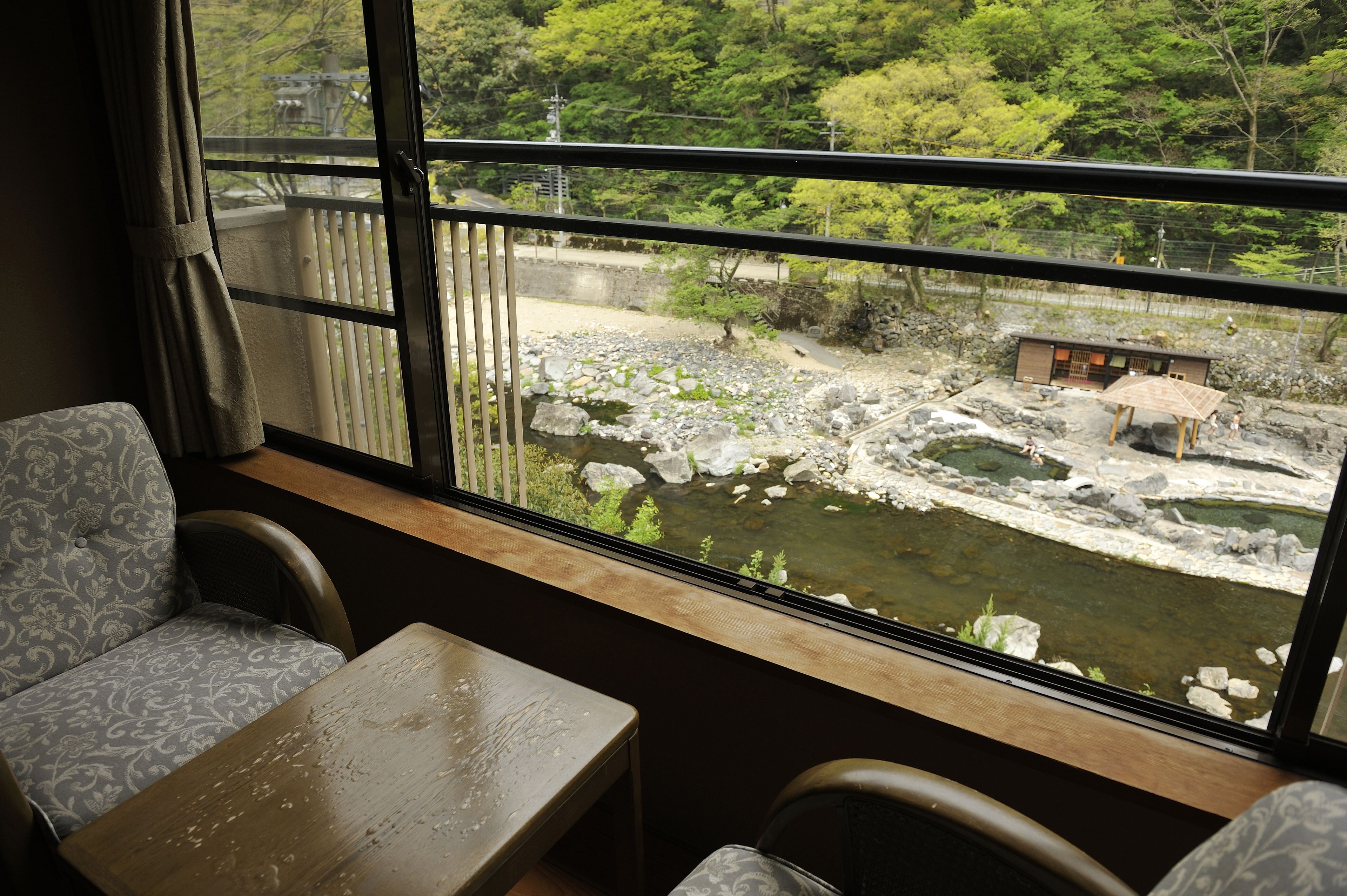 A room with a view of a rocky stream and greenery outside. There is a table and two patterned chairs.