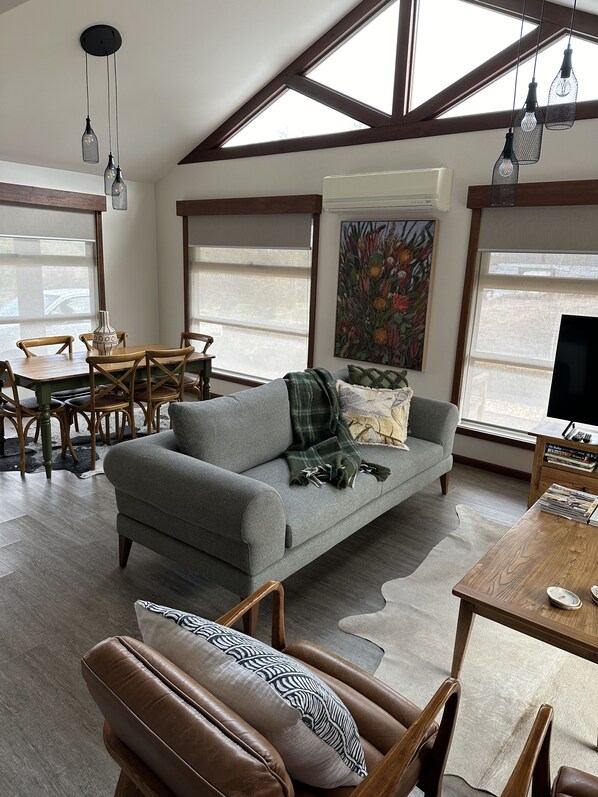 A spacious living area with a grey sofa, wooden dining table, and chairs. There is a painting on the wall, a TV on a stand, and a window with a view.