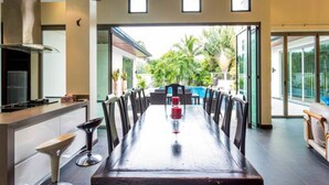 A modern dining area with a long table and chairs, a kitchen area with a stove, and a view of the outdoors. - Private Pool Villas Phuket - Mandala Arnalia (Choeng Thale)