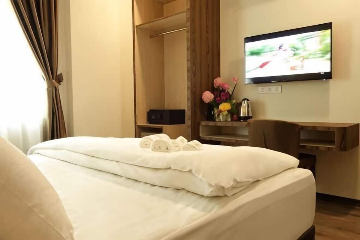 A hotel room with a bed, a television mounted on the wall, a wooden wardrobe, and a small table with a vase of flowers.