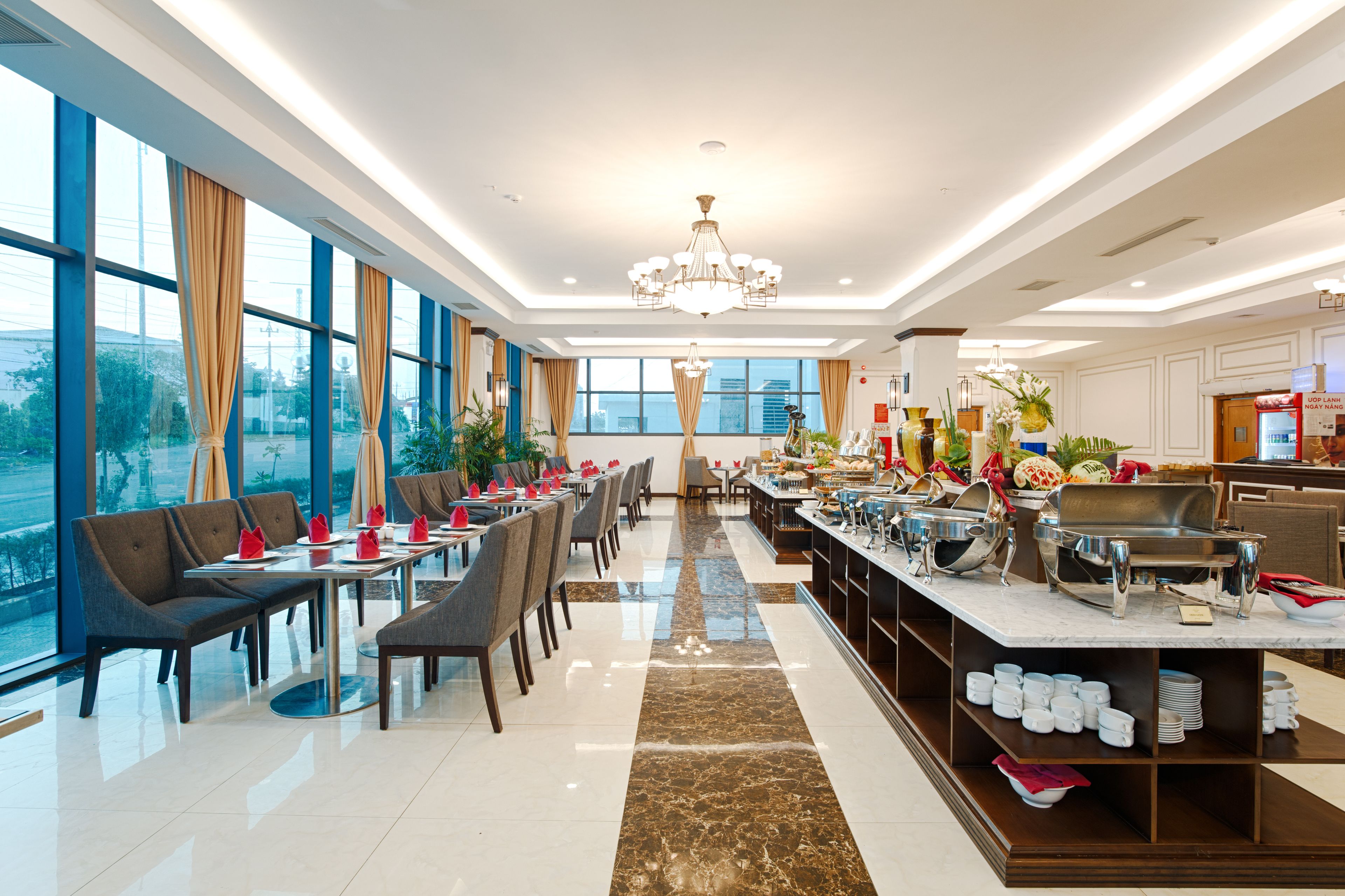 A modern hotel dining area with large windows, a central chandelier, and buffet service.