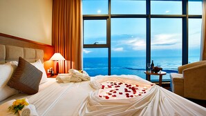 Ein Hotelzimmer mit großem Fenster, einem Bett mit weißer Bettwäsche, einem Nachttisch mit Lampe und Blick aufs Meer.