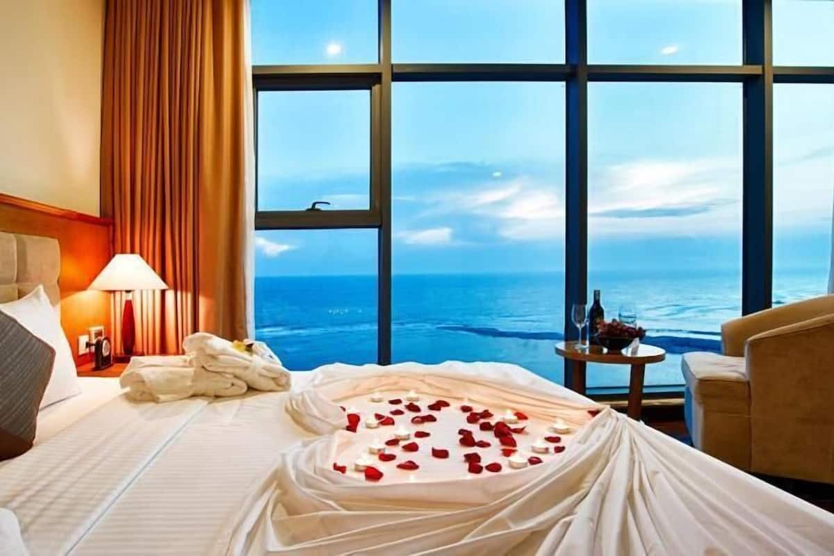 Ein Hotelzimmer mit Bett, Nachttisch, Lampe, Stuhl und Blick auf das Meer.