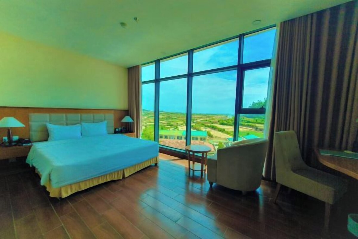Hotelkamer met een groot bed, een klein rond tafeltje, een stoel en uitzicht op een groen landschap door grote ramen.