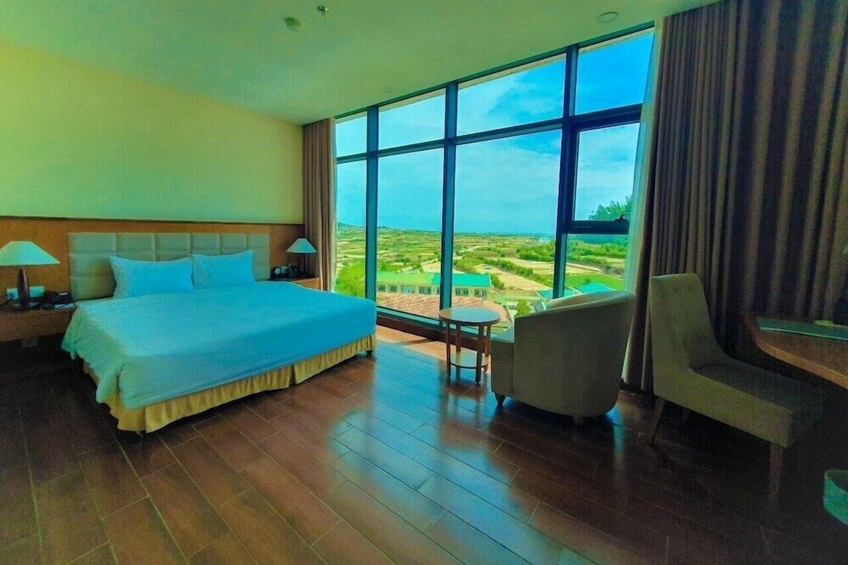 Ein Hotelzimmer mit einem großen Bett, einem Sessel, einem kleinen Tisch und Blick auf einen Golfplatz durch große Fenster.